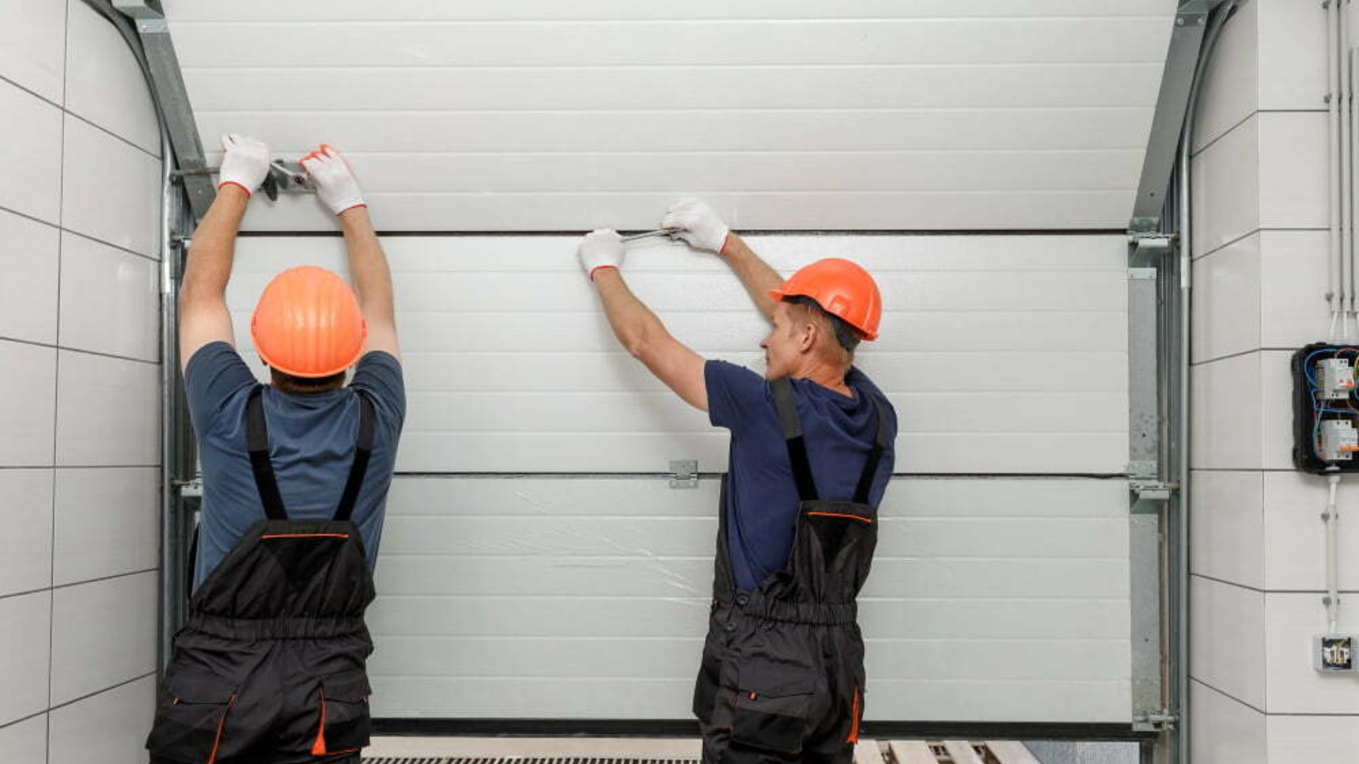 Garage door repair by two technicians wearing safety gear.